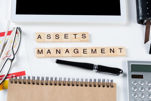 Asset management concept with letters on cubes. Still life of office workplace with supplies. Flat lay white surface with tablet computer, calculator and notepad. Investment strategy and planning.