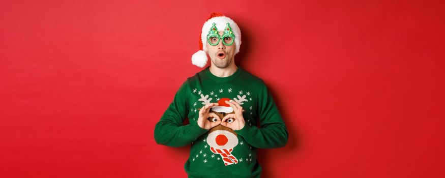 Portrait of carefree handsome man in santa hat and party glasses, making fun of his christmas sweater, looking happy over red background.