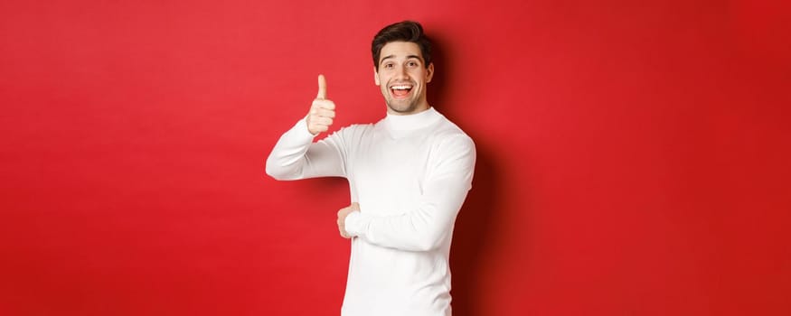 Concept of winter holidays, christmas and lifestyle. Portrait of cheerful, attractive man in white sweater, showing thumbs-up in approval, like good idea, standing over red background.