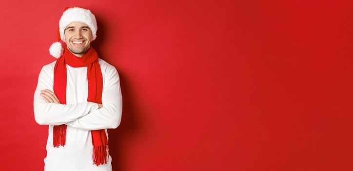 Handsome caucasian guy with bristle, wearing santa hat, scarf and white sweater, cross arms on chest and smiling happy, standing against red background.