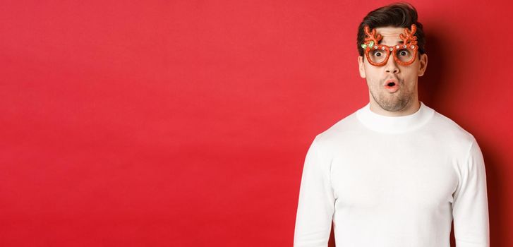 Close-up of surprised and impressed man in white sweater, party glasses, saying wow and looking amazed at camera, standing over red background.