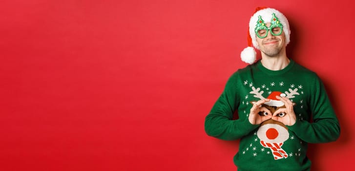 Image of happy smiling man in party glasses and santa hat, fooling around with funny christmas sweater, celebrating winter holidays, standing over red background.