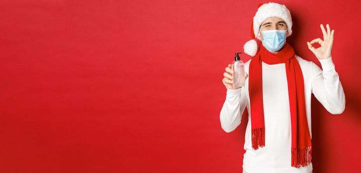 Concept of covid-19, christmas and holidays during pandemic. Cheerful man celebrating new year in medical mask and santa hat, recommending hand sanitizer, showing okay sign and winking.