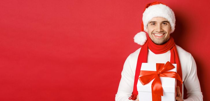 Concept of winter holidays, christmas and lifestyle. Close-up of attractive bearded guy in santa hat and scarf, holding a present, giving new year gift and smiling, red background.