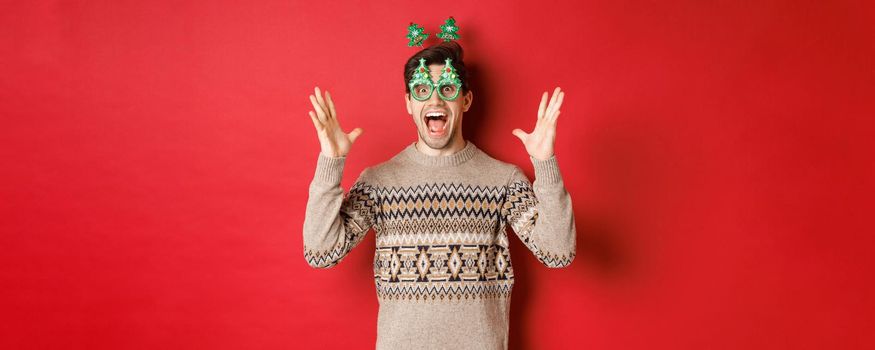 Image of excited and amazed caucasian guy in party glasses, christmas sweater, raising hands up and making big announcement, enjoying new year celebration, red background.