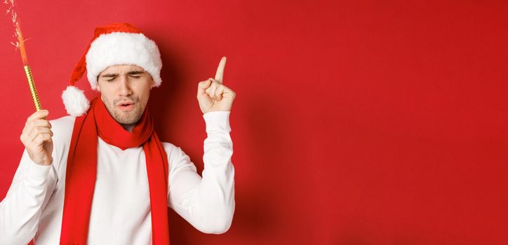 Concept of christmas, winter holidays and celebration. Attractive guy enjoying new year party, dancing with sparkler, wearing santa hat and scarf, standing over red background.