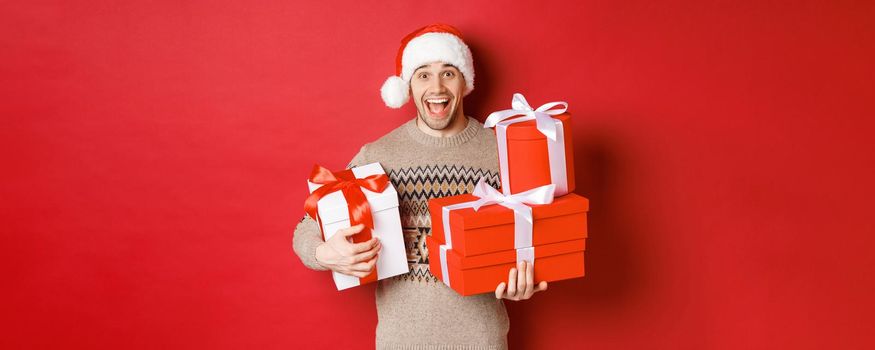 Concept of winter holidays, new year and celebration. Image of happy guy enjoying christmas, holding pile of presents and smiling amused, standing over red background.