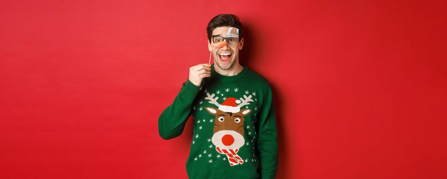 Portrait of amused handsome man in christmas sweater, holding funny party mask and smiling, celebrating winter holidays, standing over red background.
