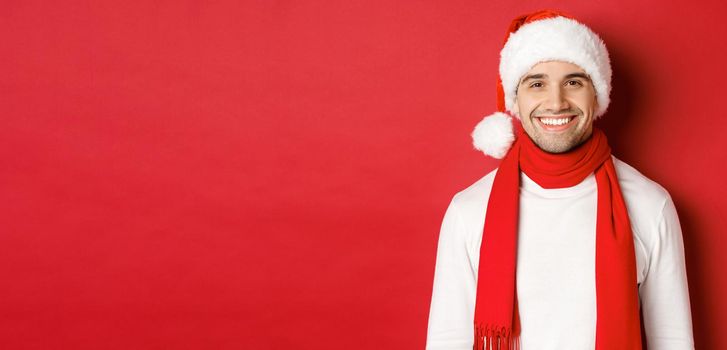 Concept of winter holidays, christmas and lifestyle. Close-up of handsome man with bristle, wearing christmas hat with scarf and smiling happy, wishing happy new year, red background.
