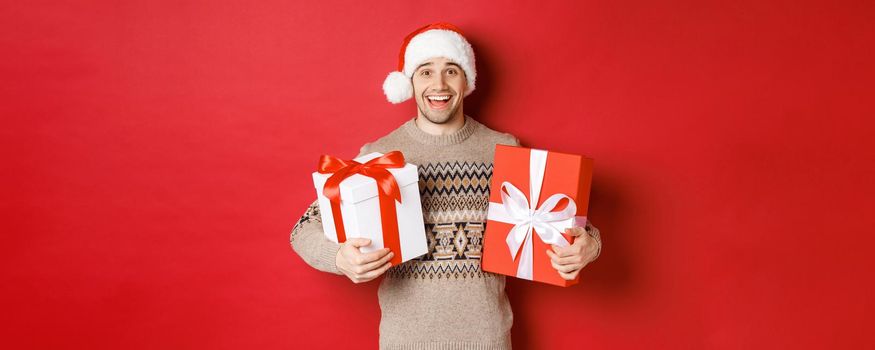 Image of cheerful attractive guy holding christmas presents, standing in santa hat and winter sweater, smiling happy, standing over red background.