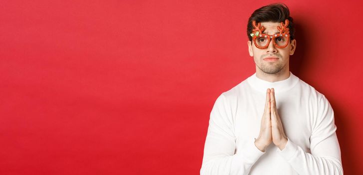 Close-up of cute guy in christmas party glasses, begging for help, need a favour, standing over red background.
