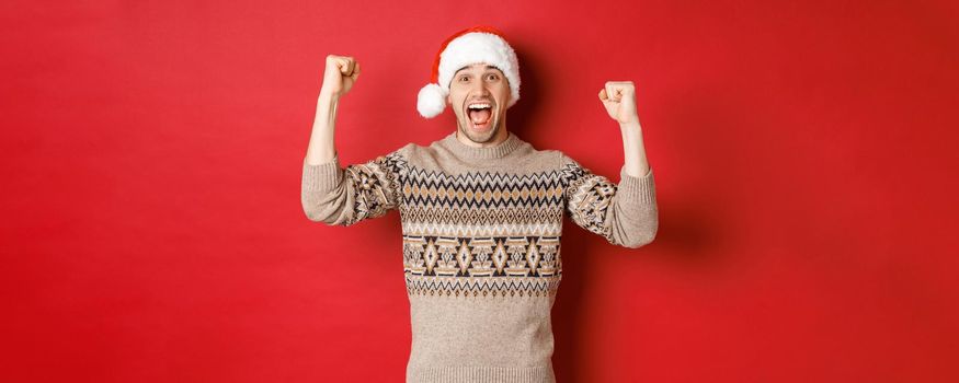 Image of cheerful handsome man in swearer and santa hat, celebrating new year, triumphing or winning something, raising hands up and shouting for joy, standing over red background.