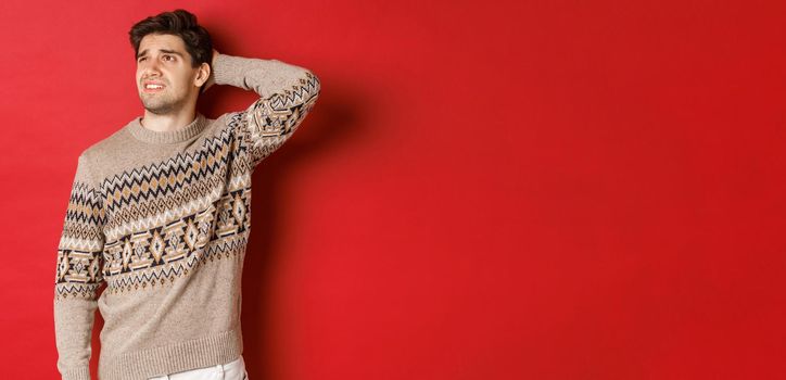 Image of puzzled and indecisive man in christmas sweater, looking at upper left corner, thinking about new year holidays gifts, standing over red background.