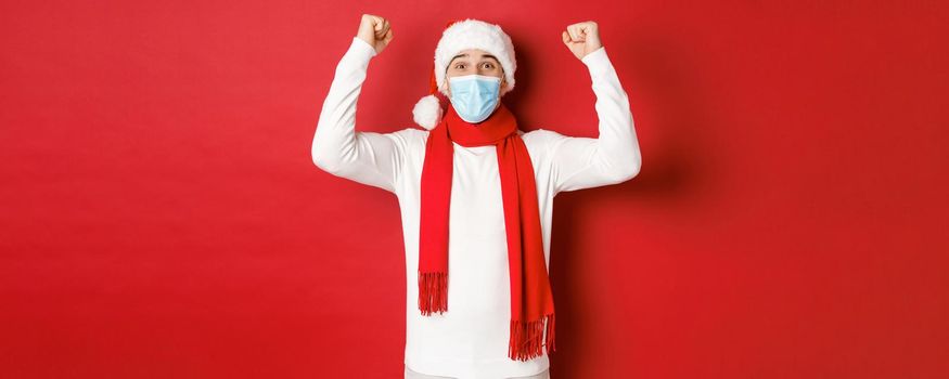 Concept of covid-19, christmas and holidays during pandemic. Portrait of happy man in santa hat and medical mask, rejoicing and celebrating new year, standing over red background.