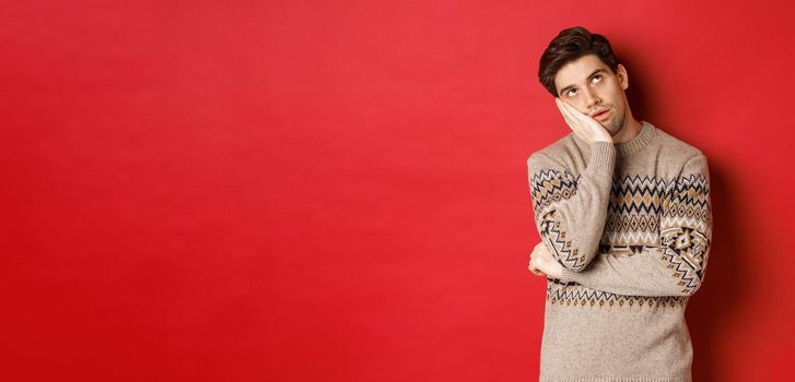 Image of annoyed young man in christmas sweater, roll eyes and leaning on hand, express displeasure and irritation, standing over red background.