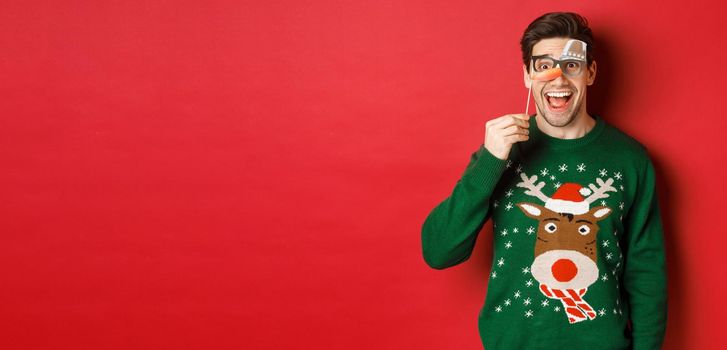 Portrait of amused handsome man in christmas sweater, holding funny party mask and smiling, celebrating winter holidays, standing over red background.