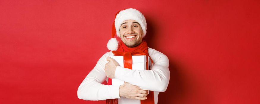 Concept of winter holidays, christmas and lifestyle. Image of lovely guy in santa hat and scarf, hugging his new year present and smiling flattered, standing over red background.