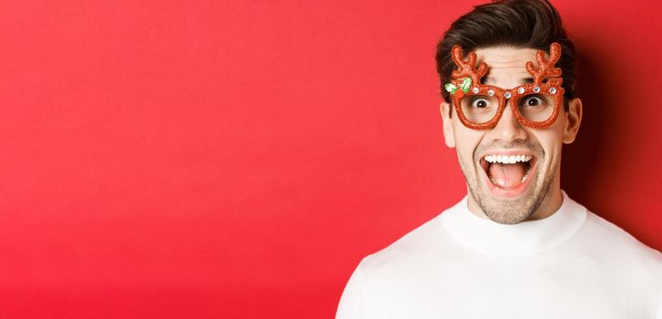 Concept of winter holidays, christmas and celebration. Close-up of attractive guy with bristle, wearing new year party glasses and looking amazed at promo offer, standing over red background.