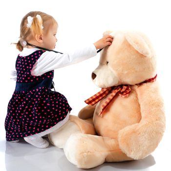 Beautiful little girl is playing doctor. She listens through a phonendoscope of a teddy bear. The concept of play and family happiness.