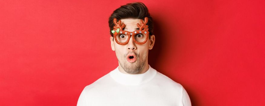 Concept of winter holidays, christmas and celebration. Close-up of attractive guy with bristle, wearing new year party glasses and looking impressed at promo offer, standing over red background.