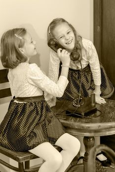 Two elegant girls sisters in beautiful dresses, talking on old phone. Black-and-white photo. Retro style.