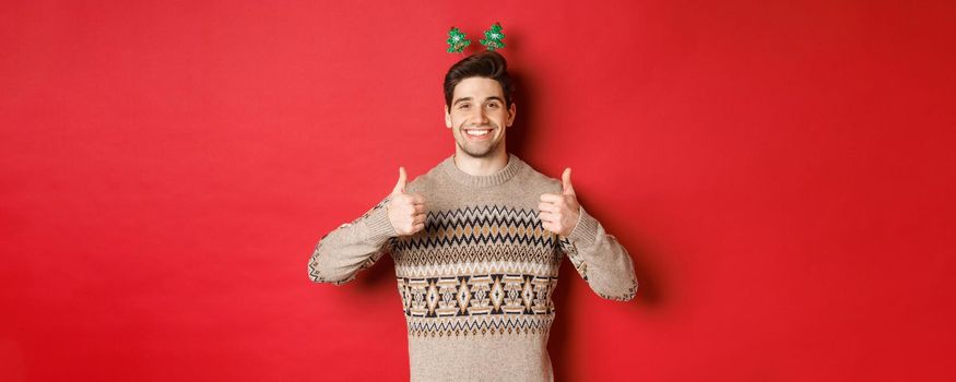 Concept of winter holidays, christmas and celebration. Cheerful bearded guy in sweater, showing thumbs-up in approval and smiling, enjoying new year party, red background.