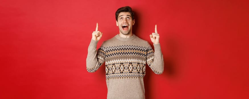 Image of excited handsome guy who likes winter holidays showing advertisement, looking and pointing fingers up with amazed face, standing in christmas sweater over red background.