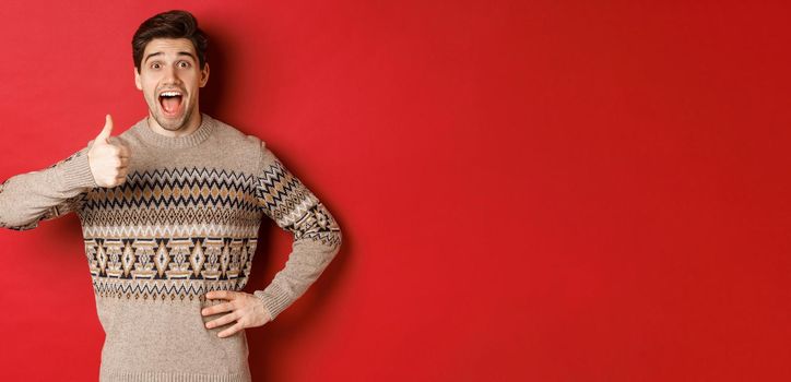 Portrait of excited, attractive man in christmas sweater, showing thumbs-up and looking amazed, recommending good thing, advertising christmas related product.