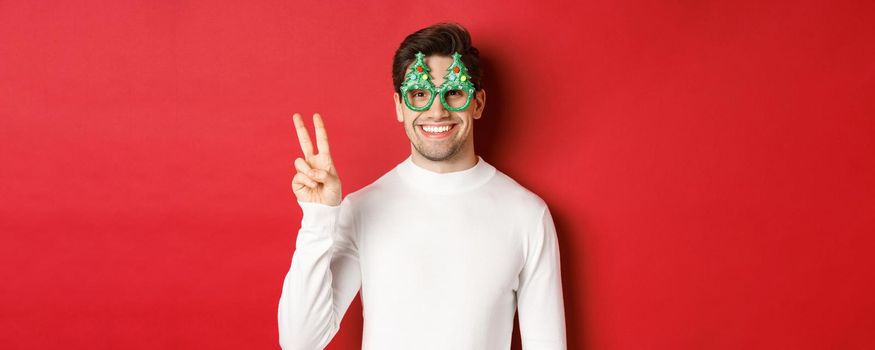 Image of handsome man in white sweater and party glasses, showing peace sign and smiling, wishing merry christmas, standing over red background.