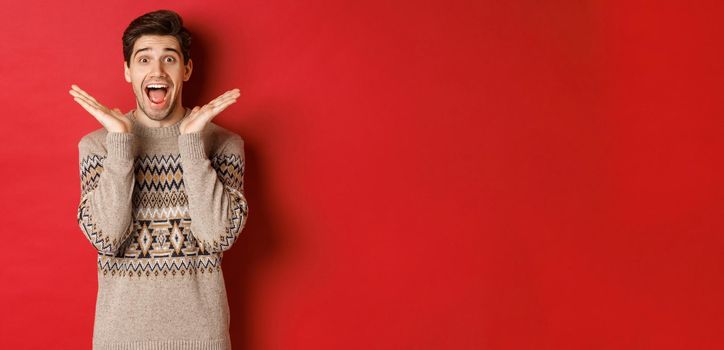 Portrait of surprised and excited guy in christmas sweater, reacting to new year present, smiling amazed, standing over red background.