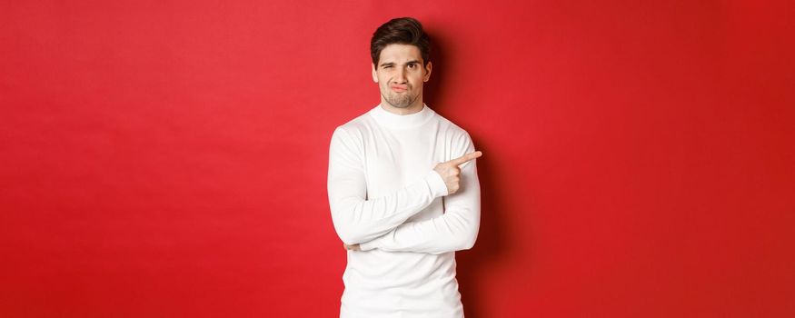 Doubtful and reluctant handsome man grimacing, pointing fingers right at something bad, standing in white sweater over red background.