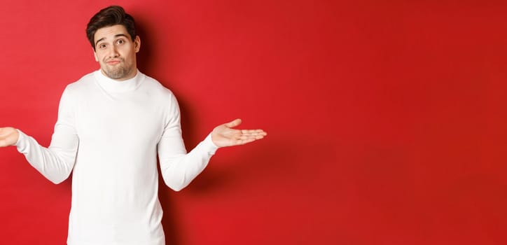 Portrait of clueless handsome guy, shrugging with hands spread sideways, being unaware, dont know anything, standing over red background.