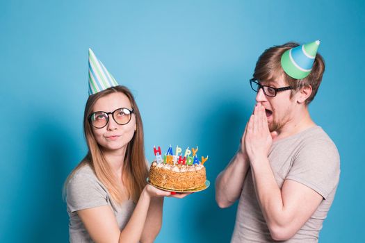 Funny young couple in paper caps and with a cake make a foolish face and wish happy birthday while standing against a blue background. Concept of congratulations and fooling around