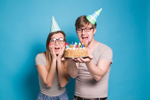 Funny young couple in paper caps and with a cake make a foolish face and wish happy birthday while standing against a blue background. Concept of congratulations and fooling around