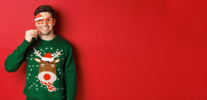 Portrait of handsome smiling man in christmas sweater and party glasses, celebrating new year and having fun, standing against red background.