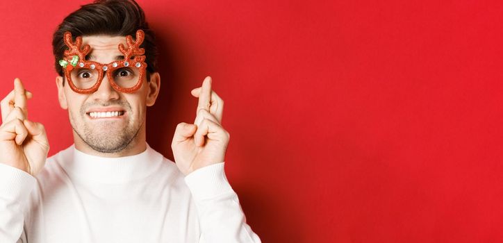 Concept of winter holidays, christmas and celebration. Close-up of nervous man in party glasses, crossing fingers for good luck and pleading, making a wish, standing over red background.
