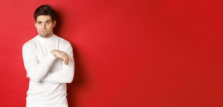 Image of sassy caucasian man in white sweater, looking thoughtful at camera, making choice, standing over red background.