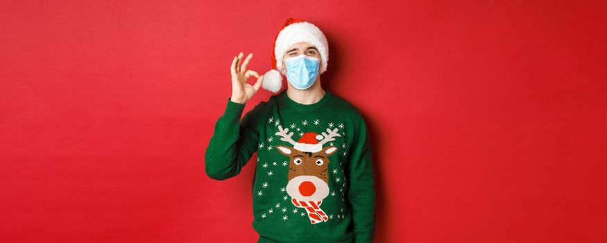 Concept of new year, covid-19 and social distancing. Cheerful man in medical mask and santa hat, showing okay sign, recommending something good, standing over red background.