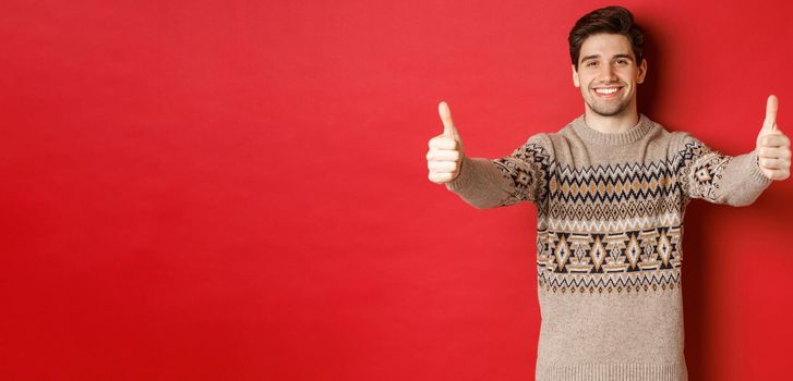 Image of handsome caucasian man in christmas sweater, showing thumbs-up in approval and smiling, wishing happy holidays, standing over red background.