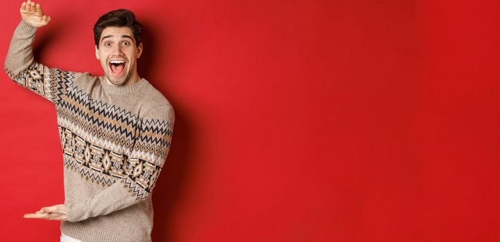 Portrait of excited and happy handsome man in christmas sweater, showing something big, holding large gift for holidays, standing over red background.