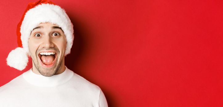Concept of winter holidays, christmas and celebration. Close-up of surprised handsome man in santa hat, hear amazing new year promo offer, standing against red background.