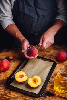Preparing of fresh and ripe peaches with honey on baking sheet. Chef halves fruit with knife.
