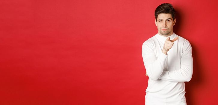 Portrait of thoughtful handsome man in white sweater, pointing at camera and making choice, standing against red background.