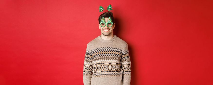 Portrait of happy and cute man in winter sweater and party glasses, celebrating new year or christmas, smiling while standing over red background.