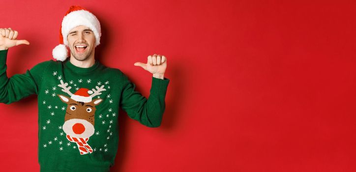 Concept of winter holidays, christmas and lifestyle. Sassy handsome man in santa hat and green sweater, pointing at himself and winking, standing over red background.