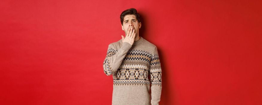 Image of tired or bored handsome man in winter sweater, yawning and covering mouth with hand, standing exhausted against red background.