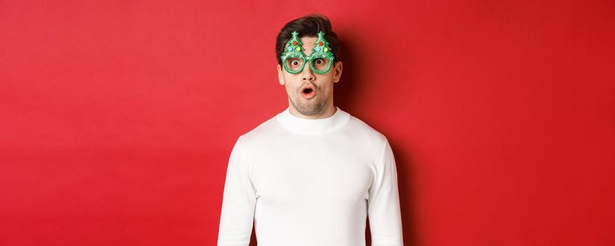 Concept of christmas, winter holidays and celebration. Close-up of surprised handsome guy in party glasses, saying wow and looking at amazing new year promo offer, red background.