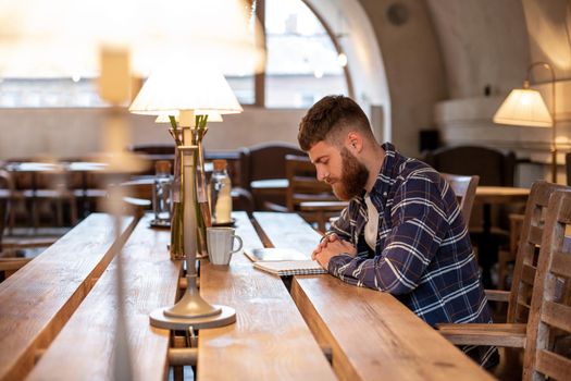Young bearded businessman sits in cafe, home at table and writes in notebook, near lies tablet computer. Man is working, studying. Online education, marketing, training.