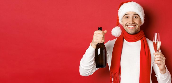 Concept of winter holidays, christmas and lifestyle. Close-up of cheerful handsome man, holding champagne bottle and glass, making toast for new year and celebrating, red background.