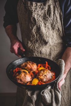 Chef holds cast iron skillet with baked chicken thighs, red bell peppers and lemon.